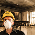 Individual with a hard hat and facemask on in front of a fire damaged kitchen.