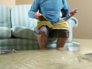 A homeowner seated on a sofa with a book on their lap, in a flooded living room while they look for ServiceMaster’s contact information on their phone